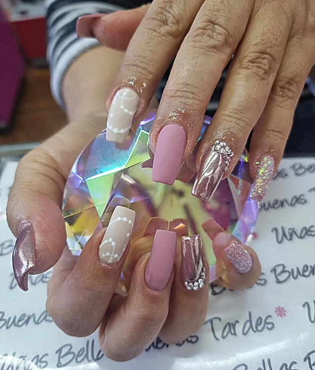 Manos sosteniendo un cristal iridiscente, con uñas largas en nude con perlitas pintadas, rosa mate, cromo rosa con pedrería y glitter