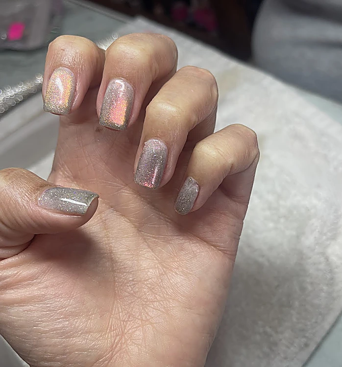 Mano con uñas cuadradas cortas de esmalte iridiscente que cambia de rosa a dorado, sobre una toalla blanca
